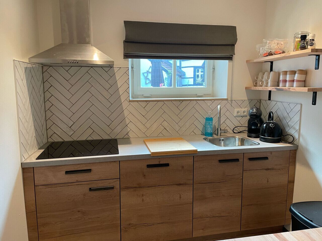 A compact kitchen with a herringbone tile backsplash, a built-in stove, a window with a view, and wooden cabinets.