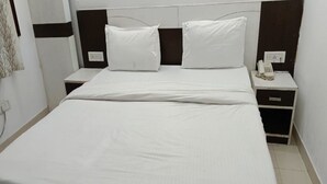 A neatly made bed with white linens, a bedside table with a telephone, and a nightstand with a lamp. - Hotel TJS Grand (New Delhi)