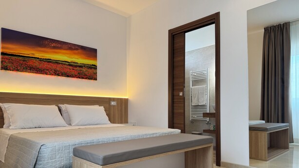 Une chambre d’hôtel moderne avec un grand lit, un banc, une salle de bain avec un miroir et un tableau représentant un coucher de soleil.