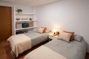 Un dormitorio con dos camas, una mesita de noche con lámpara, un estante con cajas de almacenamiento y una puerta.