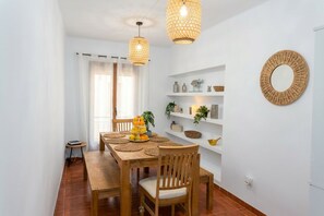 Un comedor con una mesa de madera, sillas y individuales tejidos. Hay luces colgantes, un espejo en la pared y estantes con objetos decorativos.