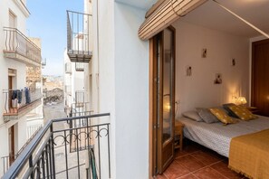 Un dormitorio con cama, mesita de noche, ventana con vista a edificios y balcón con barandillas.