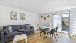 Uma sala de estar moderna com um sofá cinzento, uma mesa de centro branca e uma área de jantar com mesa redonda e cadeiras. Há duas pinturas penduradas na parede, um candeeiro de chão e uma porta de vidro deslizante para uma varanda.