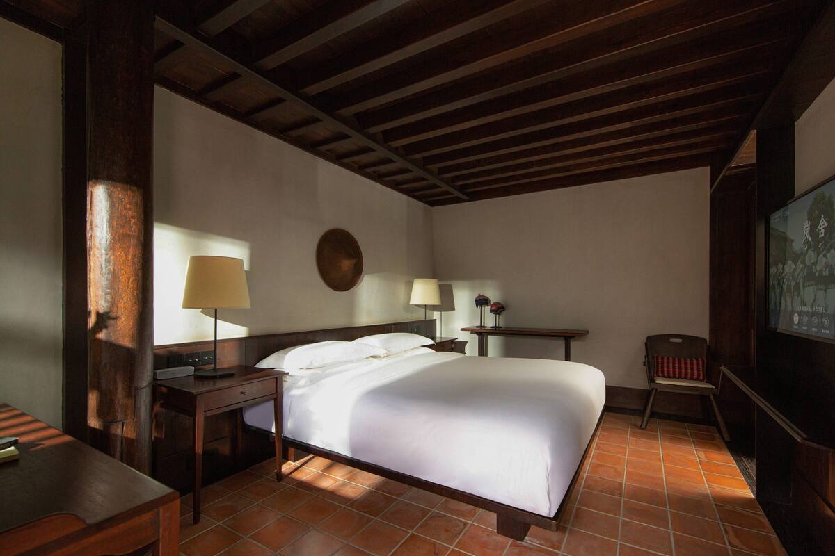 Une chambre d’hôtel avec un plafond en bois, un grand lit avec du linge de lit blanc, deux tables de chevet avec des lampes et une petite chaise.