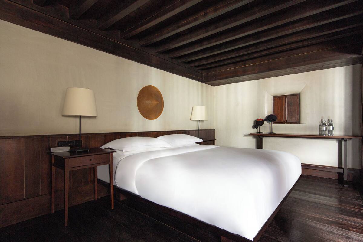 Une chambre d’hôtel avec un grand lit, deux tables de chevet, une petite table d’appoint, un miroir rond et un petit bureau avec une lampe.
