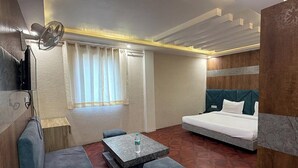 A compact hotel room with a bed, a sofa, a small table, and a window with curtains. - Hotel Beubelle (Meerut)