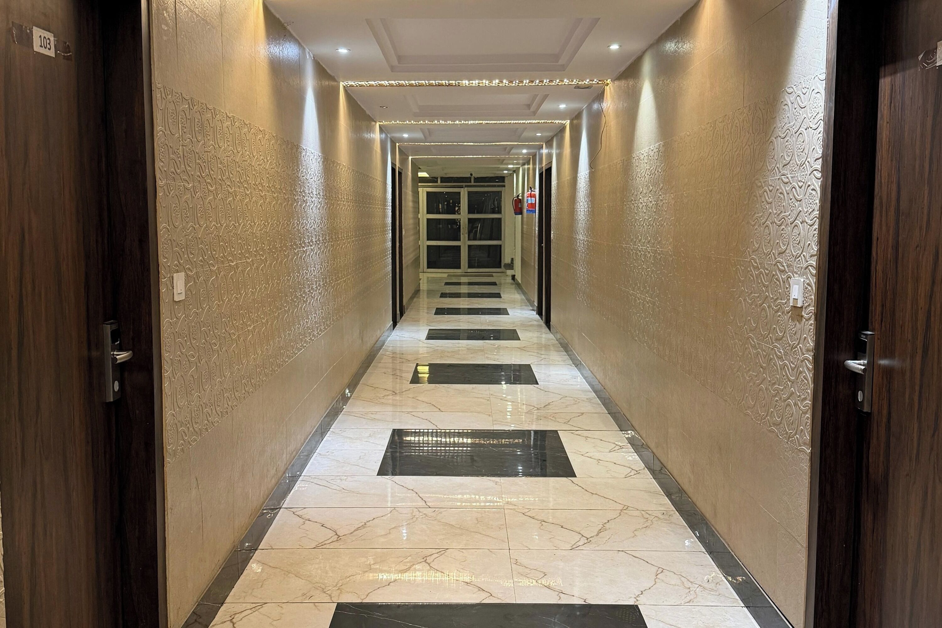 Un pasillo con paredes estampadas y piso de baldosas, que conduce a una puerta con un cartel de número.