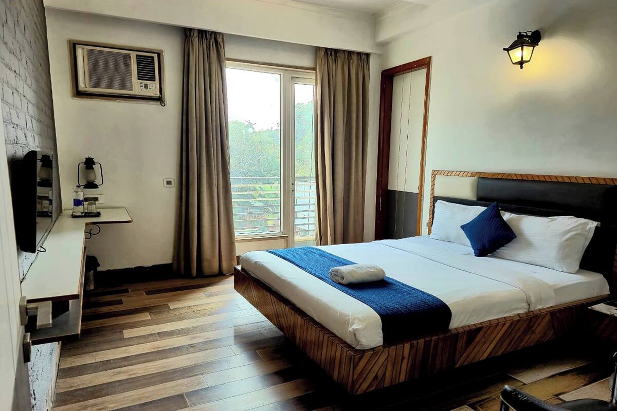 Une chambre d’hôtel avec un lit en bois, un bureau avec une lampe, une fenêtre avec des rideaux et un climatiseur fixé au mur.