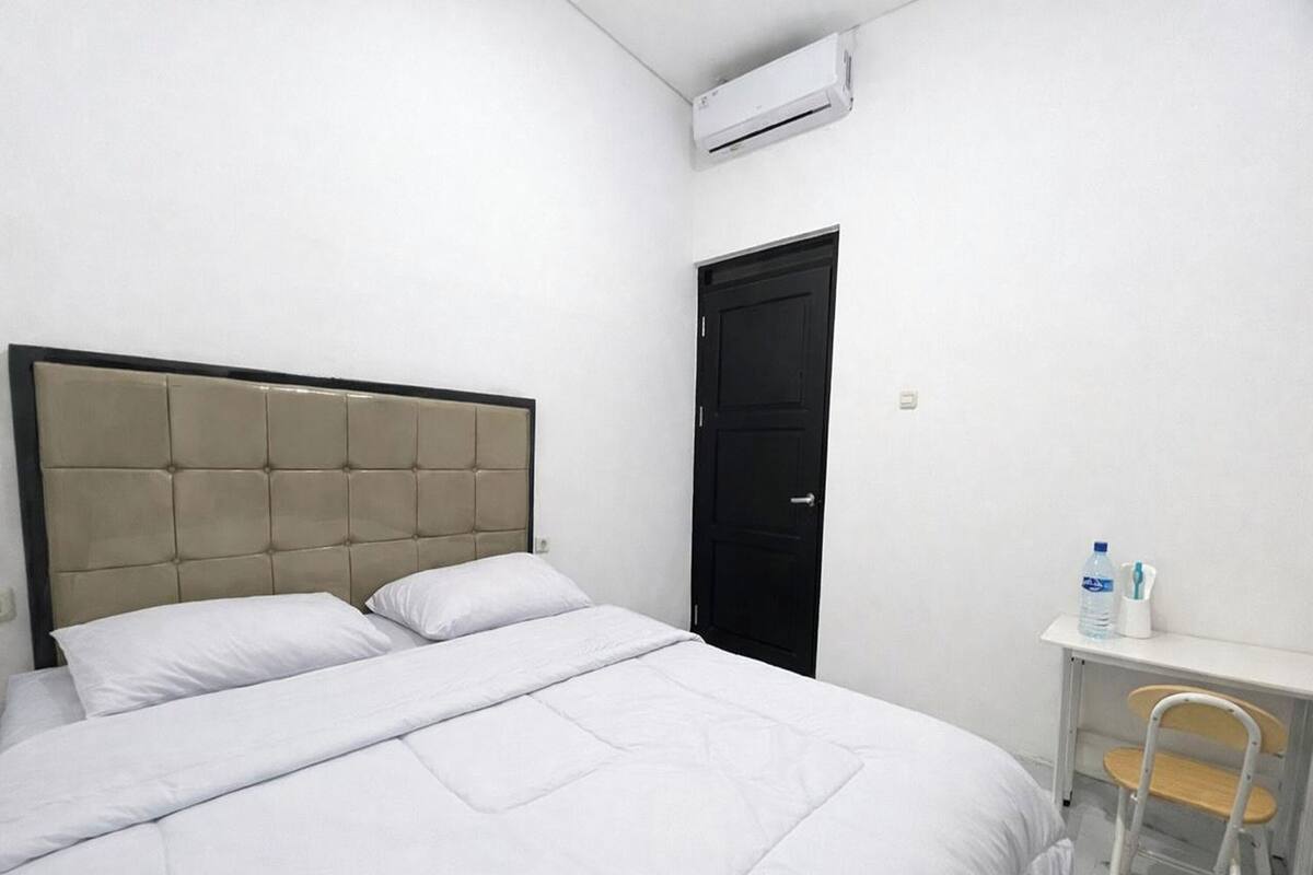 A bedroom with a bed, a desk, a chair, a door, and an air conditioning unit.
