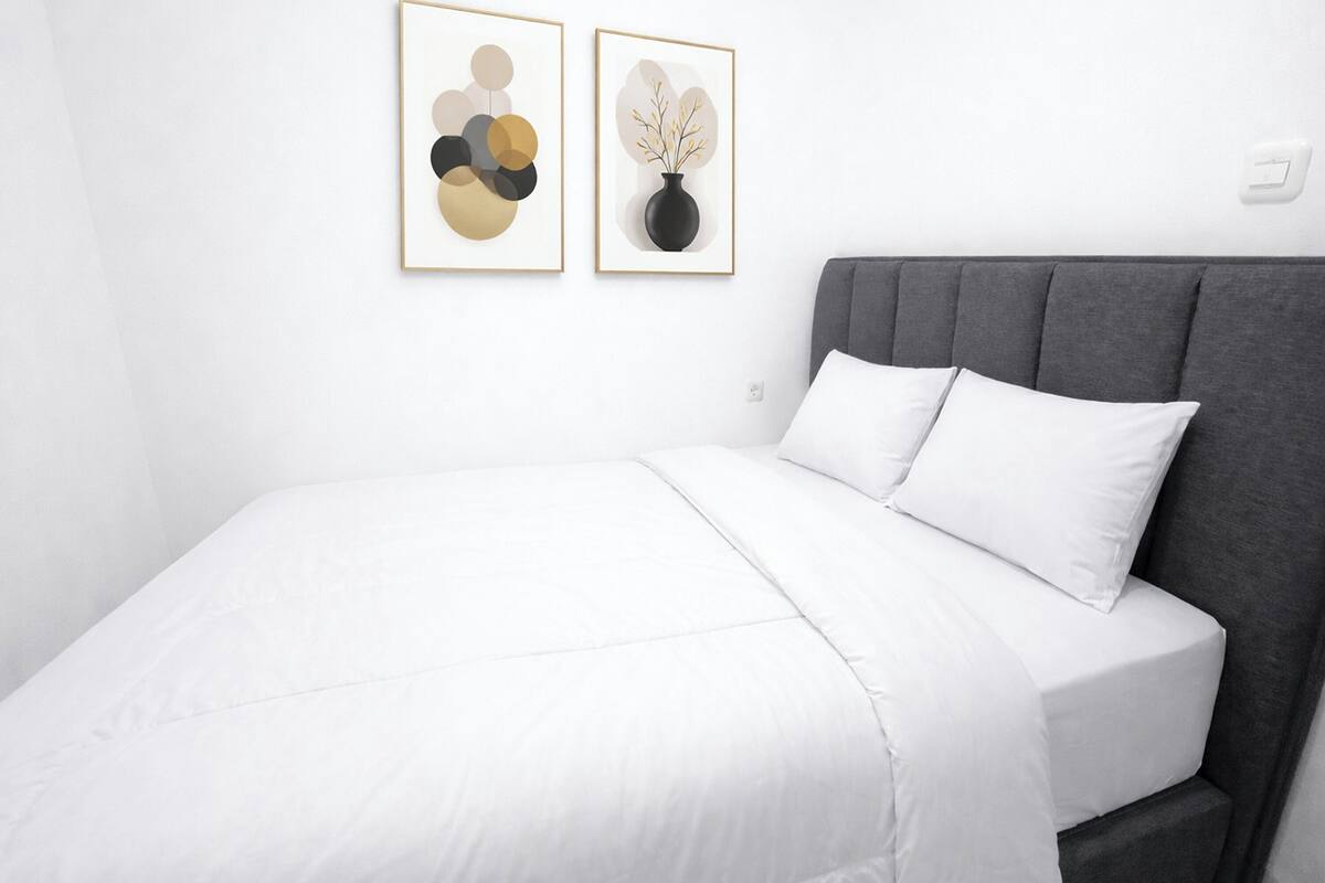 A neatly arranged bedroom with a gray headboard, white bedding, and two framed artworks on the wall.