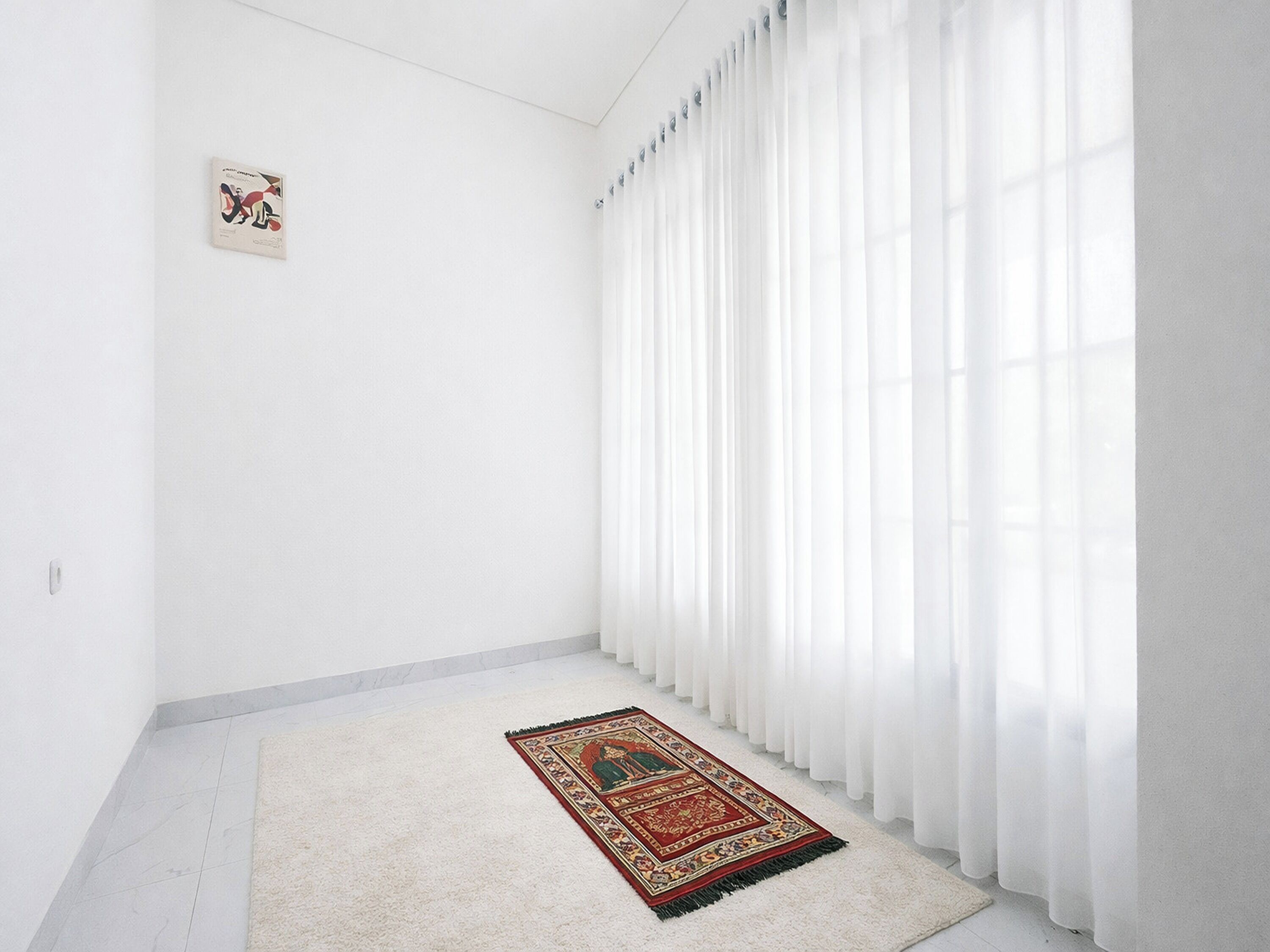 A room with white walls, a large window with sheer white curtains, a patterned rug on the floor, and a small framed picture on the wall.
