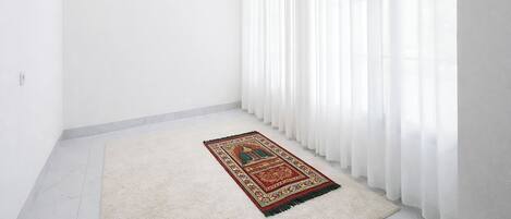 A room with white walls, a large window with sheer white curtains, a patterned rug on the floor, and a small framed picture on the wall.