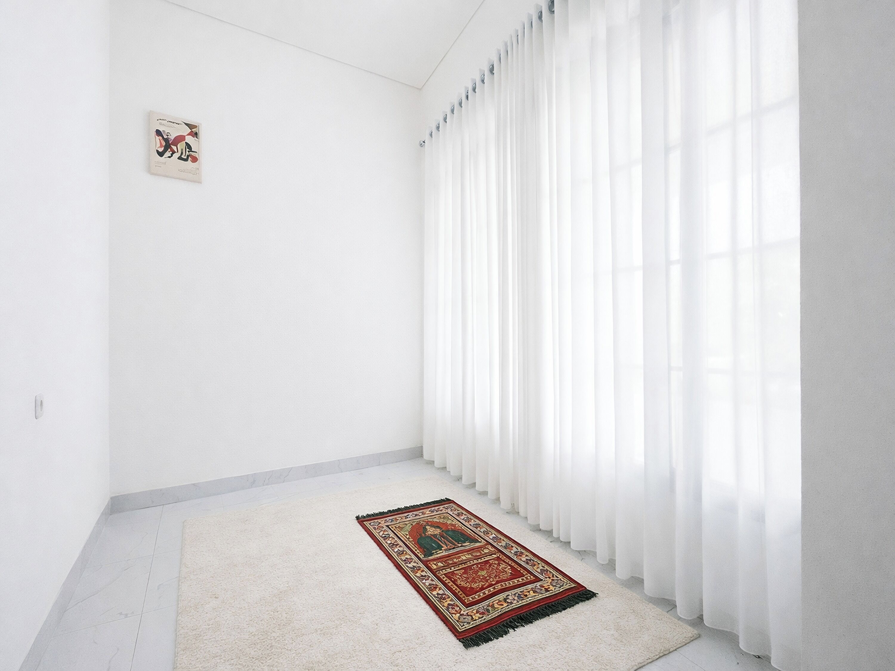 Una habitación con paredes blancas, un ventanal con cortinas blancas translúcidas, una alfombra estampada en el suelo y un pequeño cuadro en la pared.