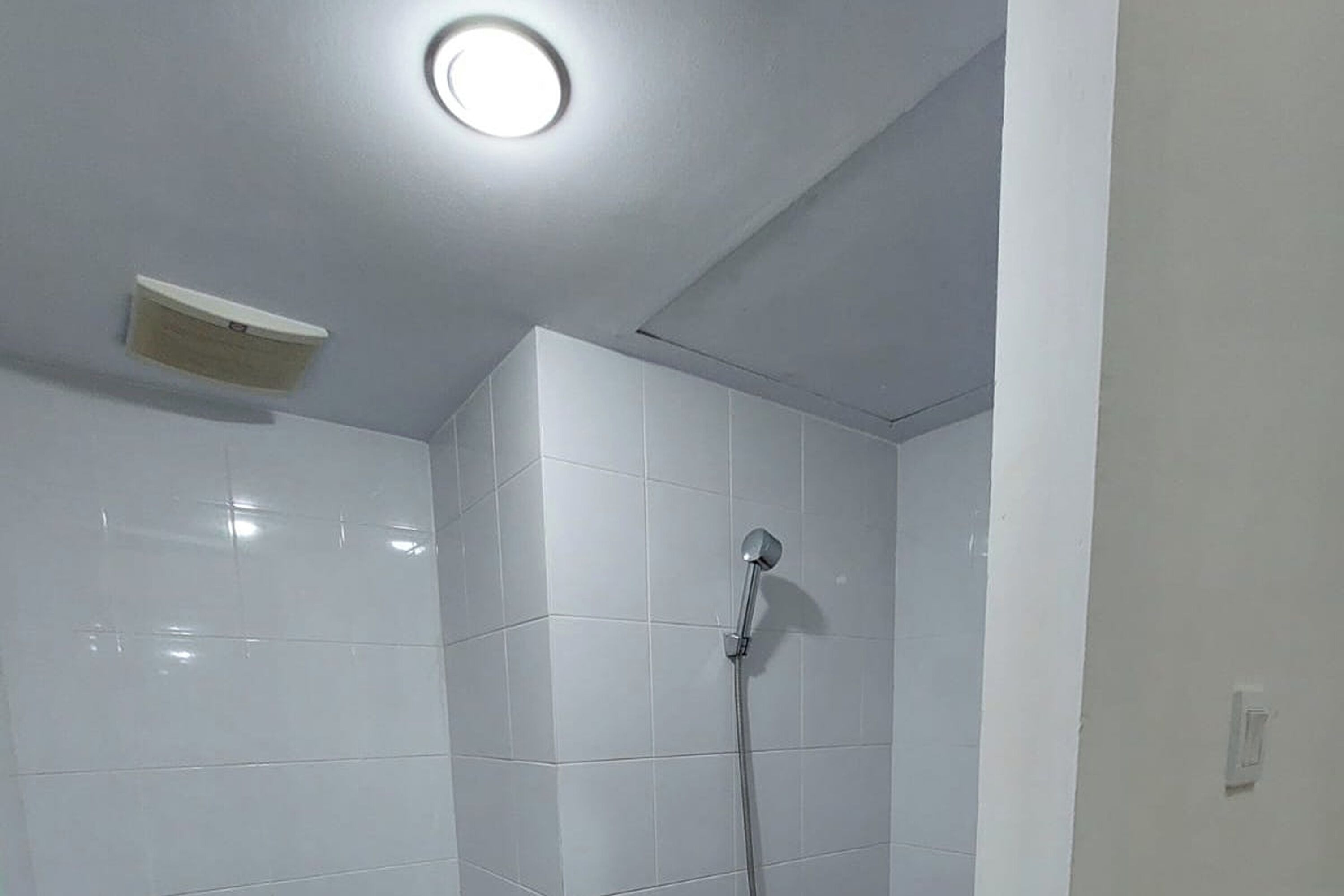 Une salle de bain avec des murs recouverts de carreaux blancs, une douche équipée d’un pommeau de douche à main et un luminaire fixé au plafond.