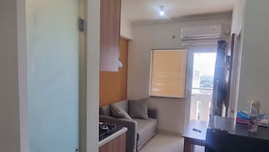 A compact living space with a kitchenette, a sofa, a window with blinds, and a ceiling-mounted air conditioner. - Apartemen Green Pramuka City By Rizal Properti (Jakarta)