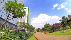 A pathway leading to a building with a green and white facade, surrounded by lush greenery and a clear sky. - Apartemen Green Pramuka City By Rizal Properti (Jakarta)