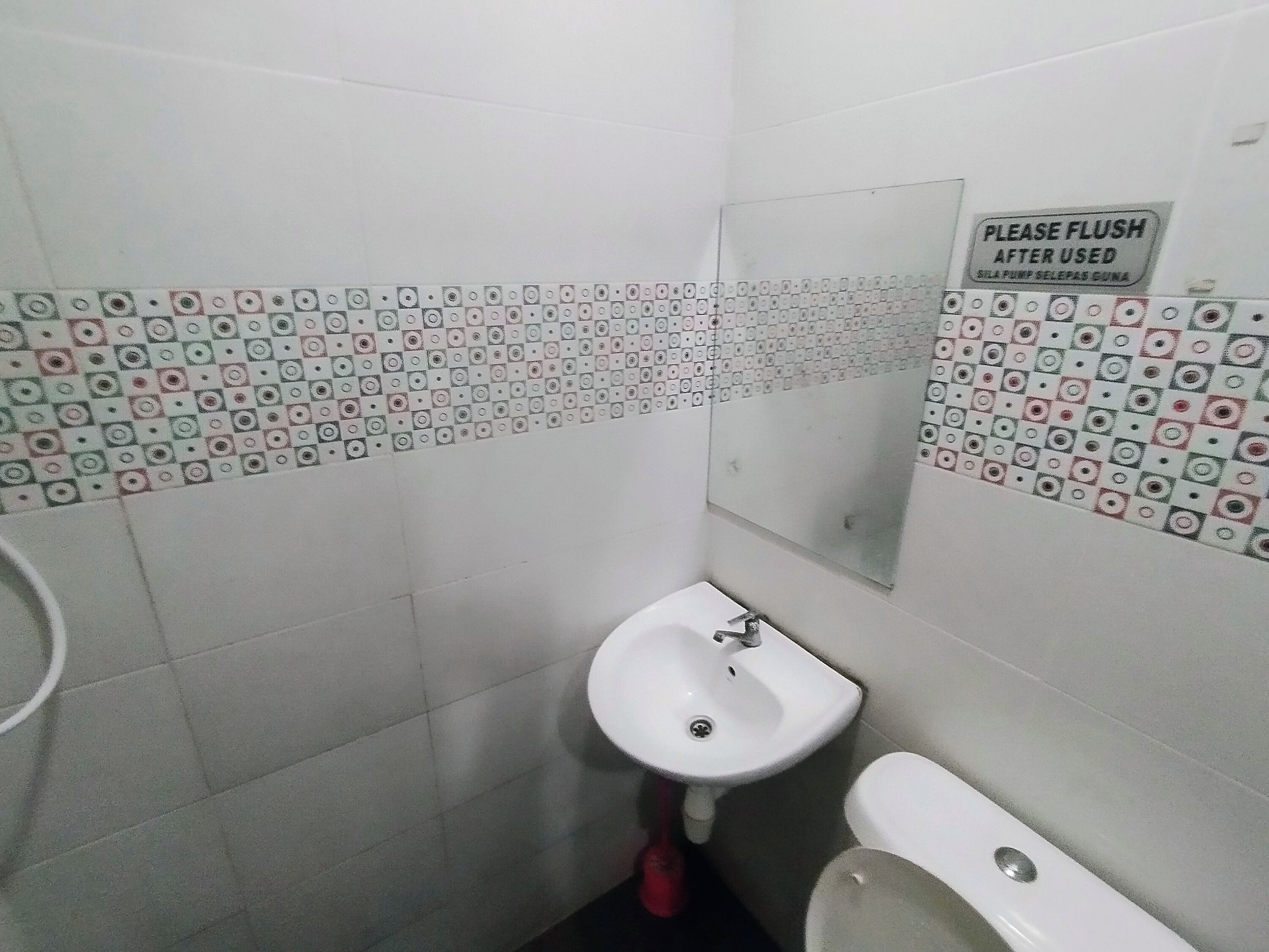 A small bathroom with a white sink, a toilet, and a mirror. The walls are tiled with a pattern of squares and circles. There is a sign on the wall that says "PLEASE FLUSH AFTER USED".