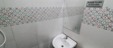 A small bathroom with a white sink, a toilet, and a mirror. The walls are tiled with a pattern of squares and circles. There is a sign on the wall that says "PLEASE FLUSH AFTER USED".