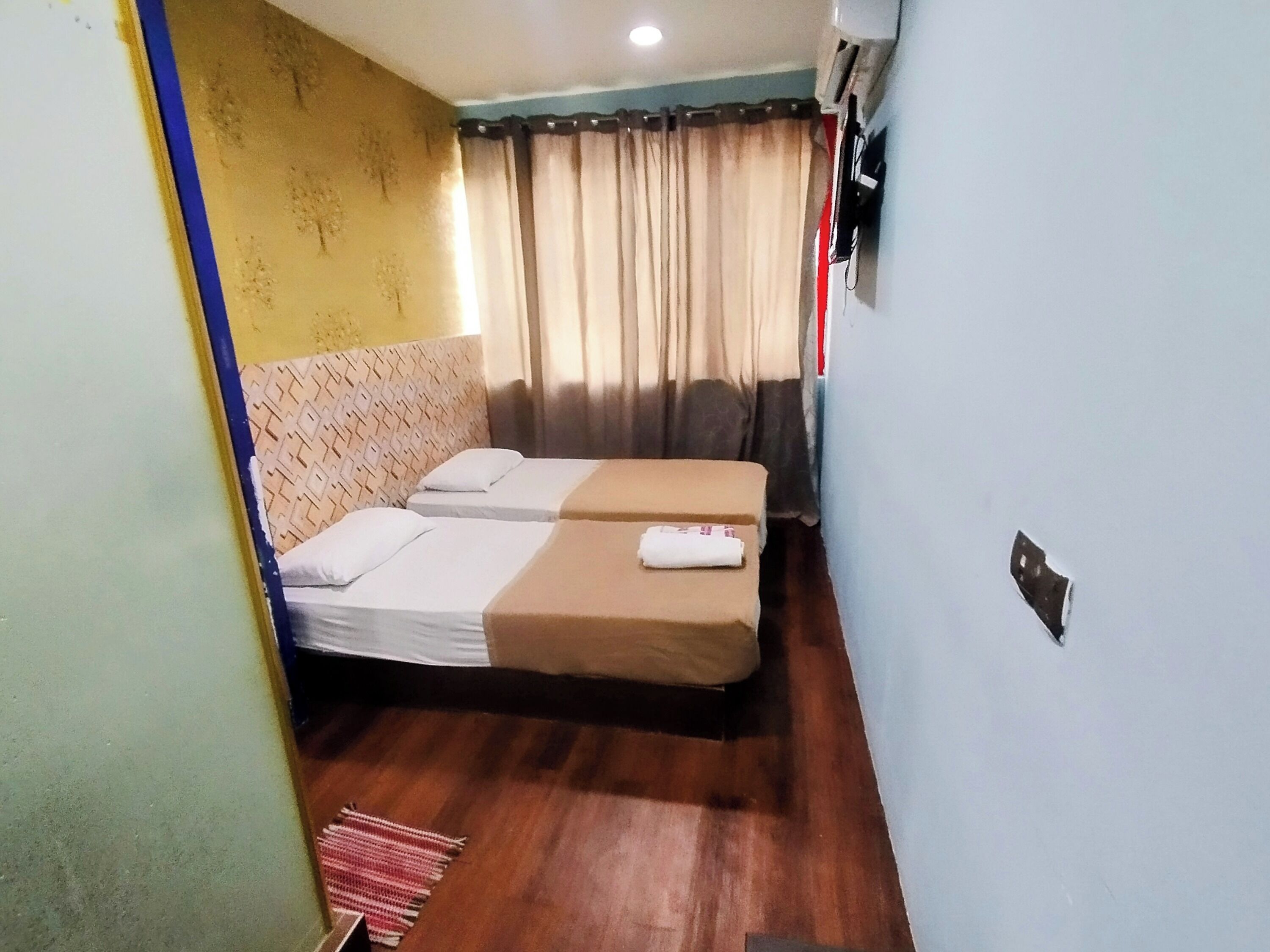 A small hotel room with two single beds, a patterned headboard, a wooden floor, and a window with curtains.