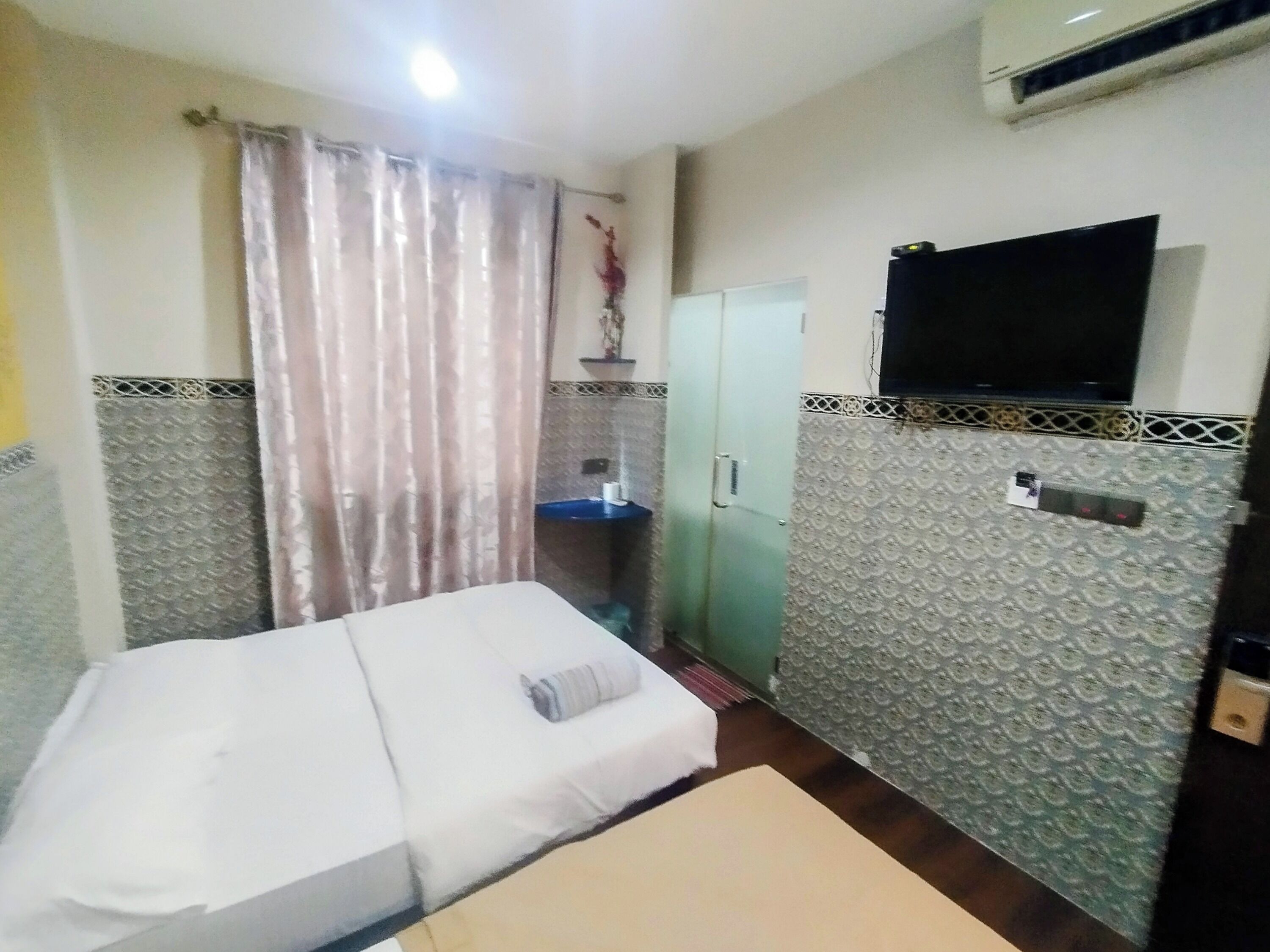 Une petite chambre avec un lit simple, un téléviseur fixé au mur, une armoire et un petit coin lavabo.