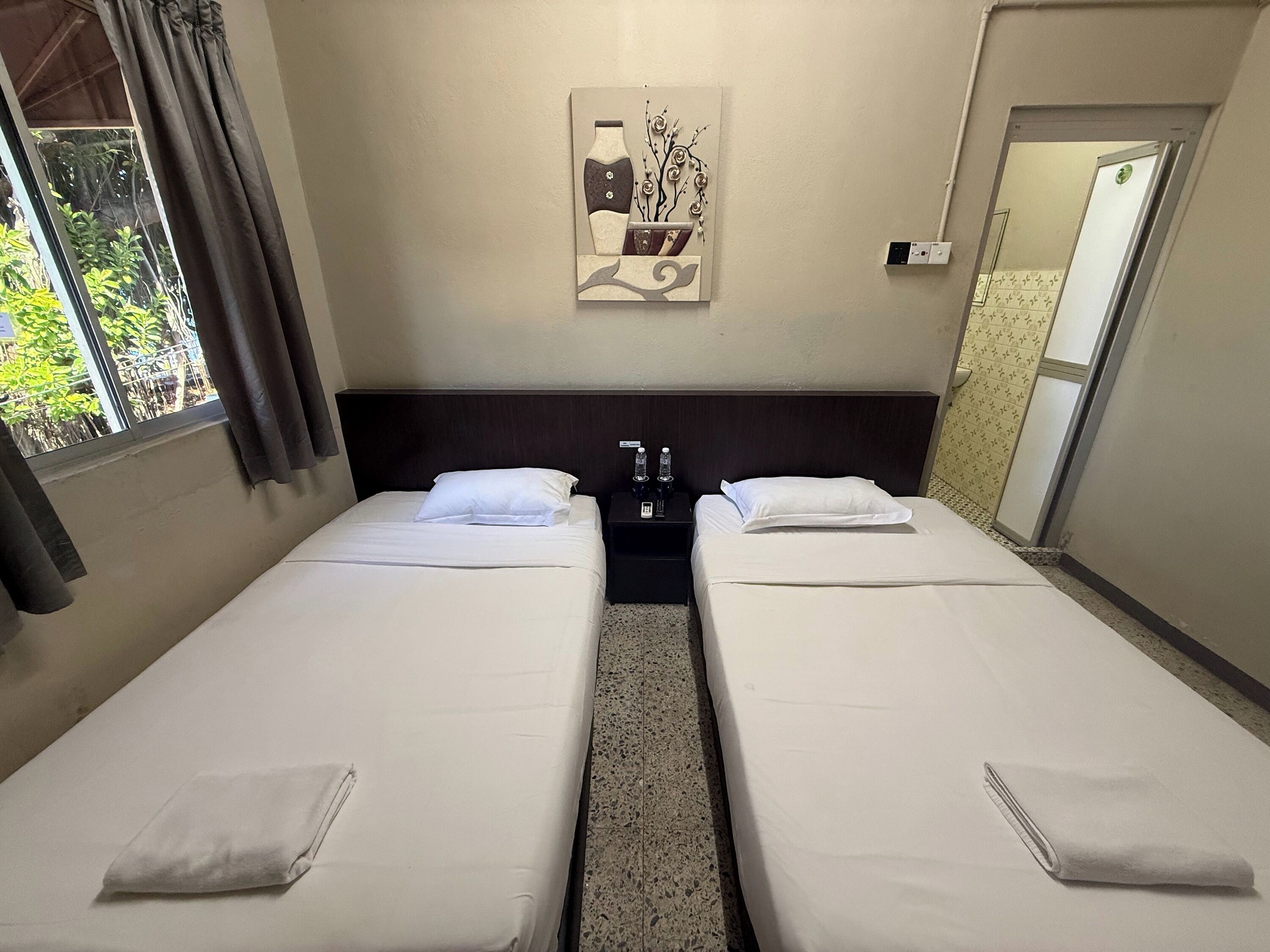 A hotel room with two single beds, a nightstand with a phone and water bottle, a wall-mounted artwork, and a window with curtains.