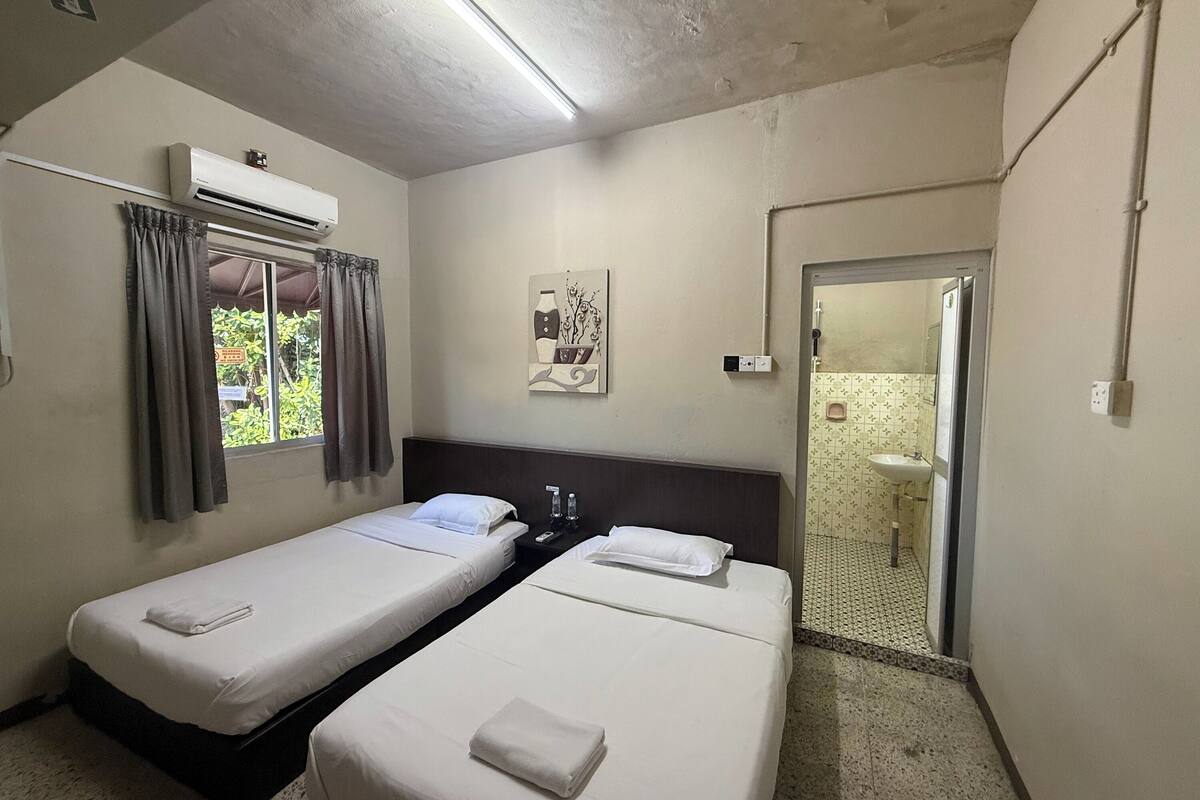 A room with two beds, an air conditioning unit, a window with curtains, a painting on the wall, and a bathroom visible through an open door.