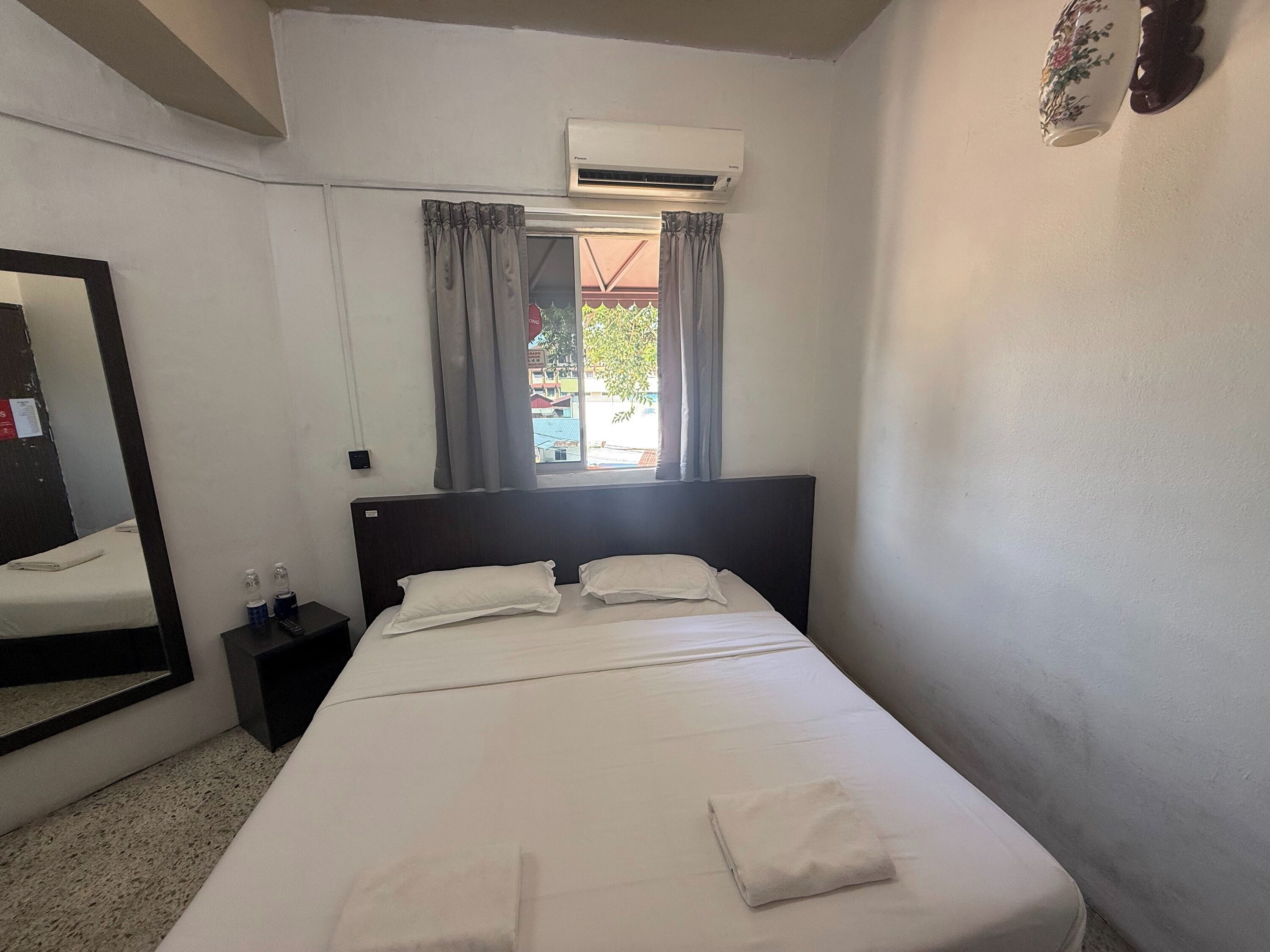 A hotel room with a bed, a nightstand, an air conditioning unit, a window with curtains, and a mirror.