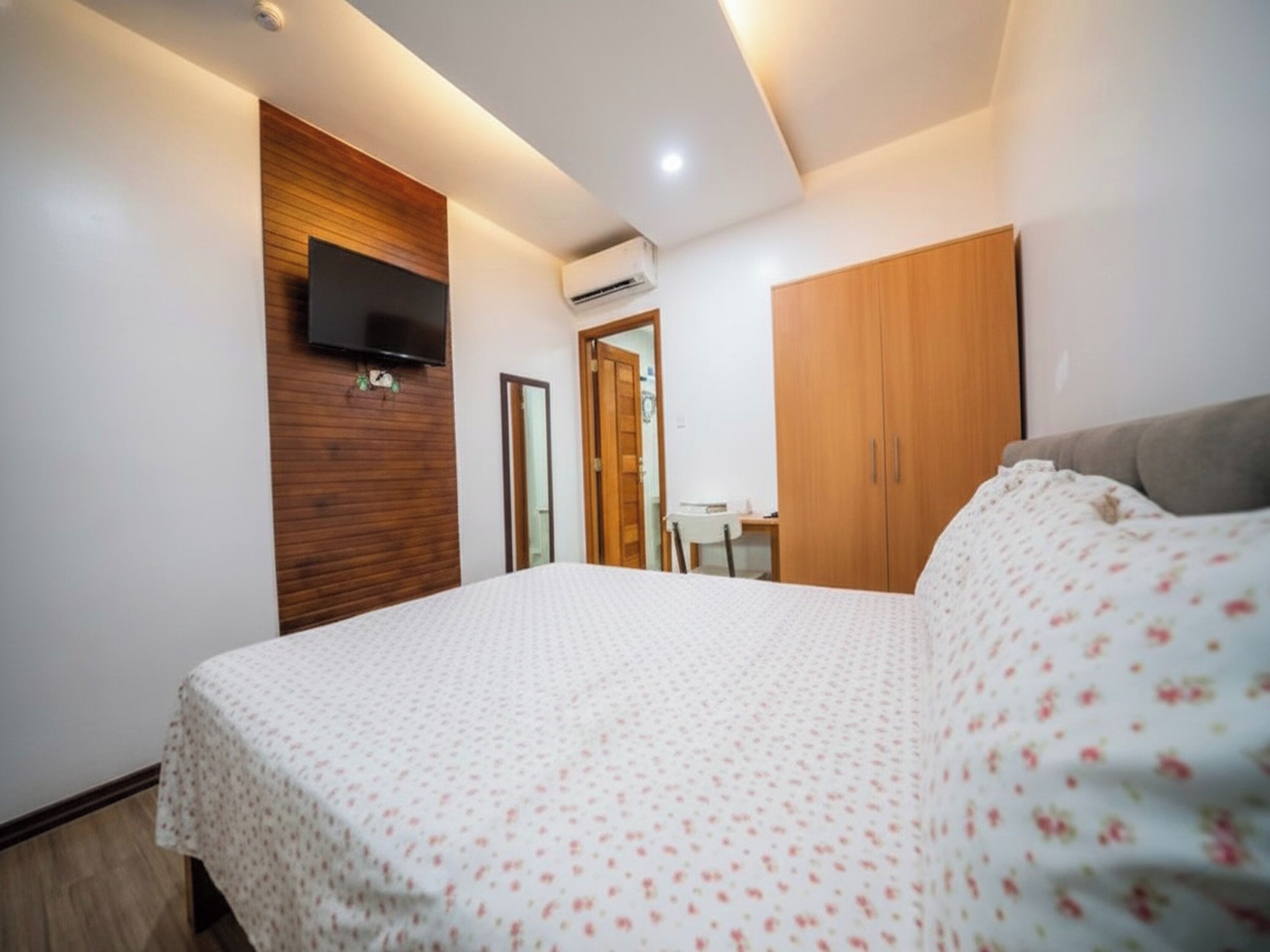 A bedroom with a bed, a TV mounted on the wall, a wardrobe, and a desk with a chair.