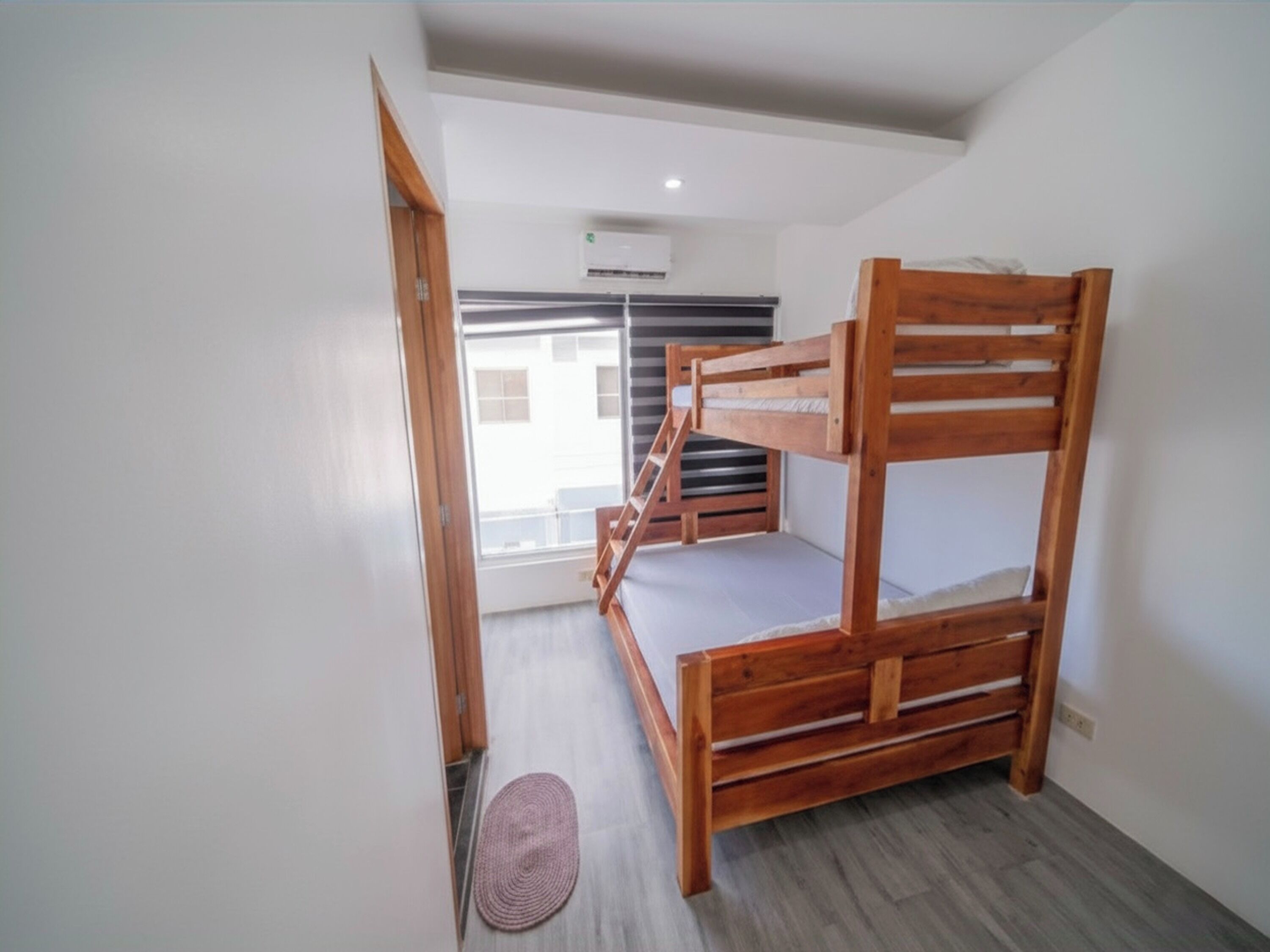A room with a bunk bed, a window with blinds, an air conditioning unit, and a wooden door.