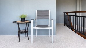 Una silla blanca y una mesita redonda con una planta en maceta en un balcón con pared gris y baranda de madera.