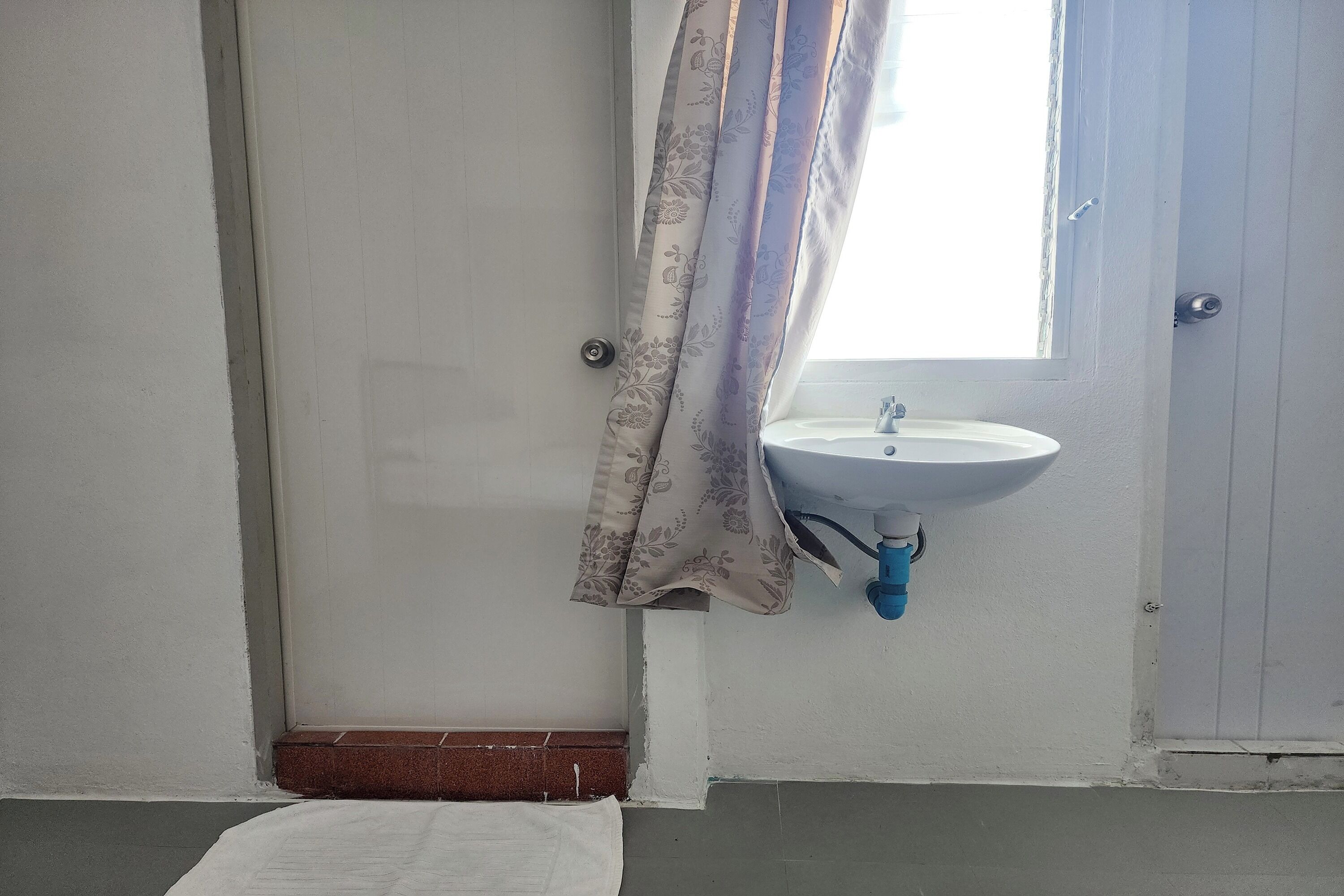 A bathroom with a white sink, a window with a curtain, and a white towel on the floor.