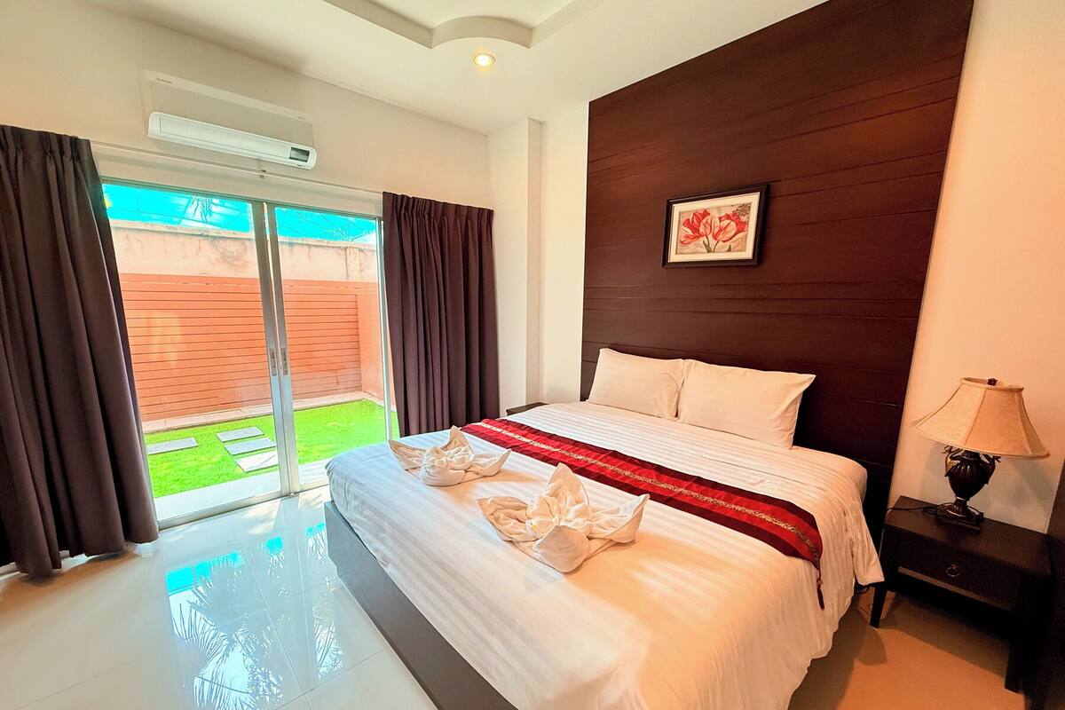 A hotel room with a large bed, a view of a green garden through a sliding glass door, a wooden headboard, and a nightstand with a lamp.