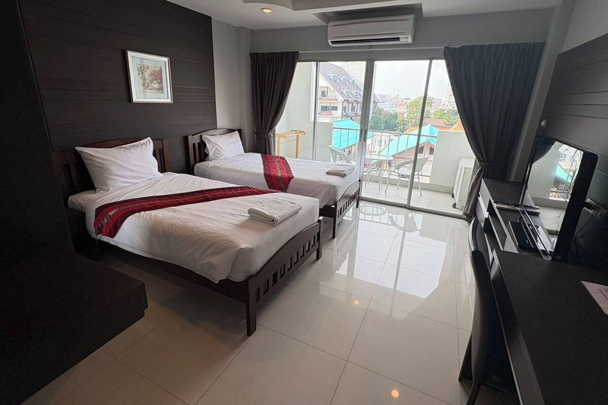 A hotel room with two beds, a TV, and a balcony with a view of buildings.