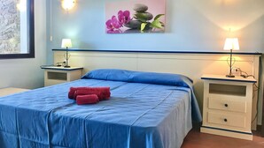 A hotel room with a bed, two bedside tables with lamps, a wall-mounted picture, and a window with a stone wall view. - Functional 28 sq m room.Comprising a double bed, two single beds, and a private bathroom with shower, bidet, and hairdryer. The room also features an outdoor space with a sea view, a patio or terrace, and a table and chairs. Breakfast is included in the  (Stintino)