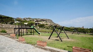 A playground with swings and a slide, surrounded by stone pathways and terraced houses on a hillside. - Functional 28 sq m room.Comprising a double bed, two single beds, and a private bathroom with shower, bidet, and hairdryer. The room also features an outdoor space with a sea view, a patio or terrace, and a table and chairs. Breakfast is included in the  (Stintino)