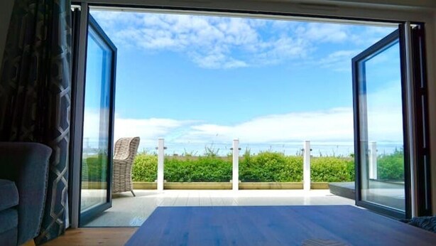 A room with a large sliding glass door leading to a balcony with a view of the sea and greenery. - Holywell Luxury Lodge with Hot Tub | Relax, Unwind & Enjoy Cornish Sunsets (Goonhavern)
