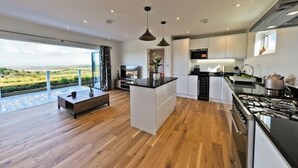 A modern kitchen with a large island, wooden flooring, and a view of the countryside. - Holywell Luxury Lodge with Hot Tub | Relax, Unwind & Enjoy Cornish Sunsets (Goonhavern)