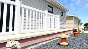 A mobile home with a white porch and a small garden area with potted plants and a mushroom-shaped planter. - Holywell Luxury Lodge with Hot Tub | Relax, Unwind & Enjoy Cornish Sunsets (Goonhavern)