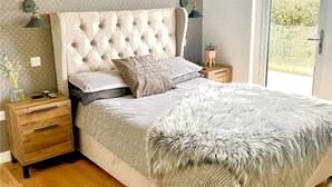 A bedroom with a tufted headboard, a fluffy throw blanket, a wooden nightstand with drawers, and a small potted plant. - Holywell Luxury Lodge with Hot Tub | Relax, Unwind & Enjoy Cornish Sunsets (Goonhavern)