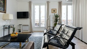 A modern living room with a black sofa, a TV, a dining table with fruit, and a balcony with curtains. - Luxembourg Gardens Cosy Studio - Paris 5th (Paris)
