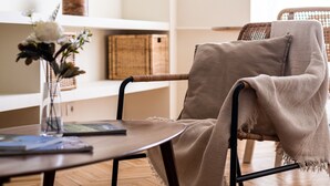 Une chaise en osier avec un plaid beige, une table ronde ornée d’un vase de fleurs et d’un livre, et des paniers en osier posés sur une étagère.