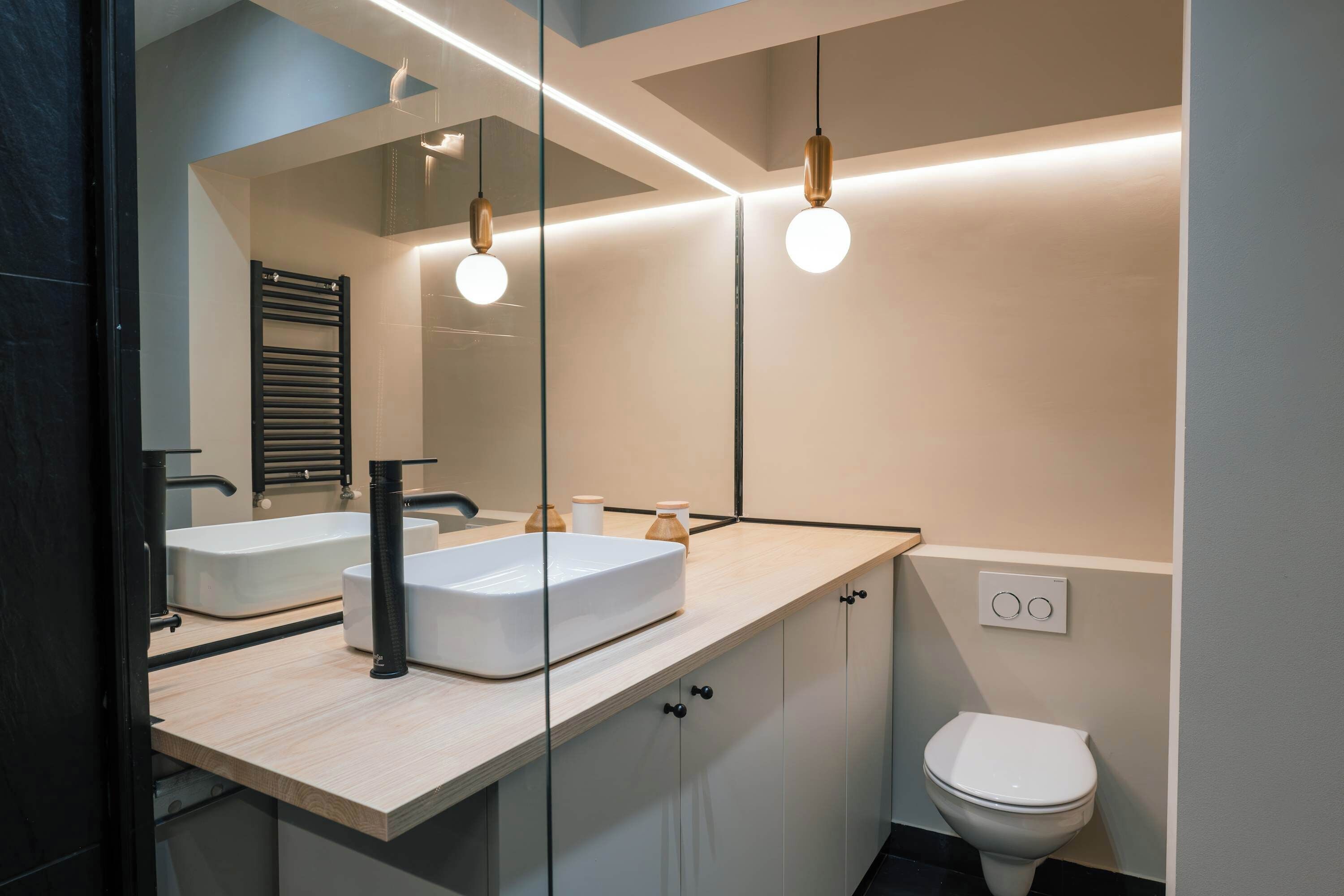Un baño moderno con un espejo grande, dos lavamanos, un inodoro y luces colgantes.