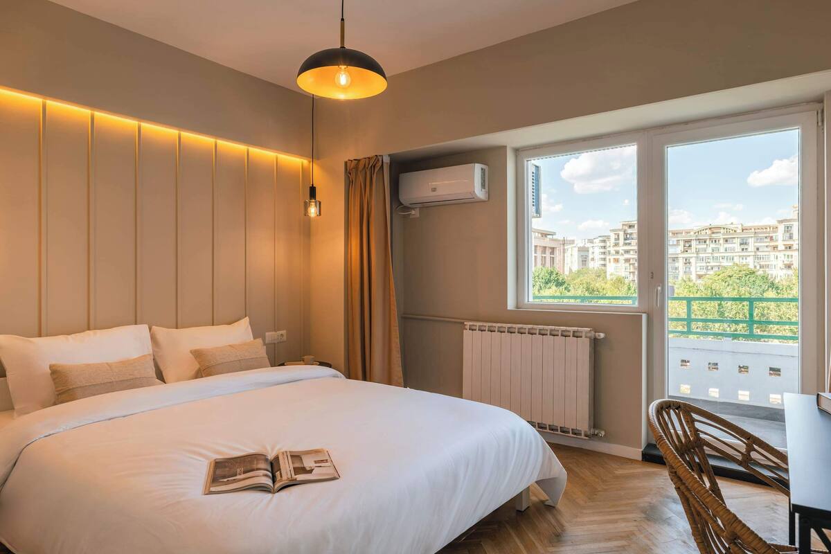 Une chambre avec un lit, un livre, une fenêtre donnant sur des immeubles, un climatiseur et une table avec une chaise.