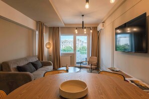 Un salon moderne avec un canapé, une table à manger agrémentée d’un saladier, un téléviseur et une vue sur les immeubles par la fenêtre.