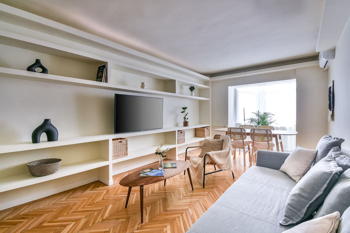 Un salon moderne avec un canapé, un téléviseur, un parquet en bois disposé en chevrons, des étagères blanches et un coin repas comprenant des chaises et une table.