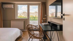 Une chambre d’hôtel équipée d’un lit, d’un bureau, d’une chaise, d’une télévision et d’une fenêtre donnant sur des immeubles.