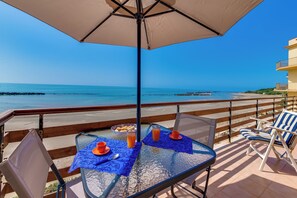 Un balcon avec une table en verre, deux chaises et un parasol. La vue comprend une plage et un ciel dégagé.