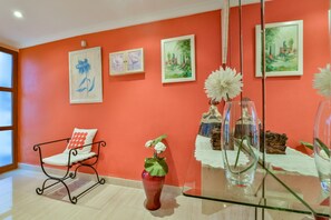 Une pièce avec un mur rouge, une chaise blanche avec un coussin rouge, un vase rouge contenant des fleurs blanches et des tableaux encadrés.