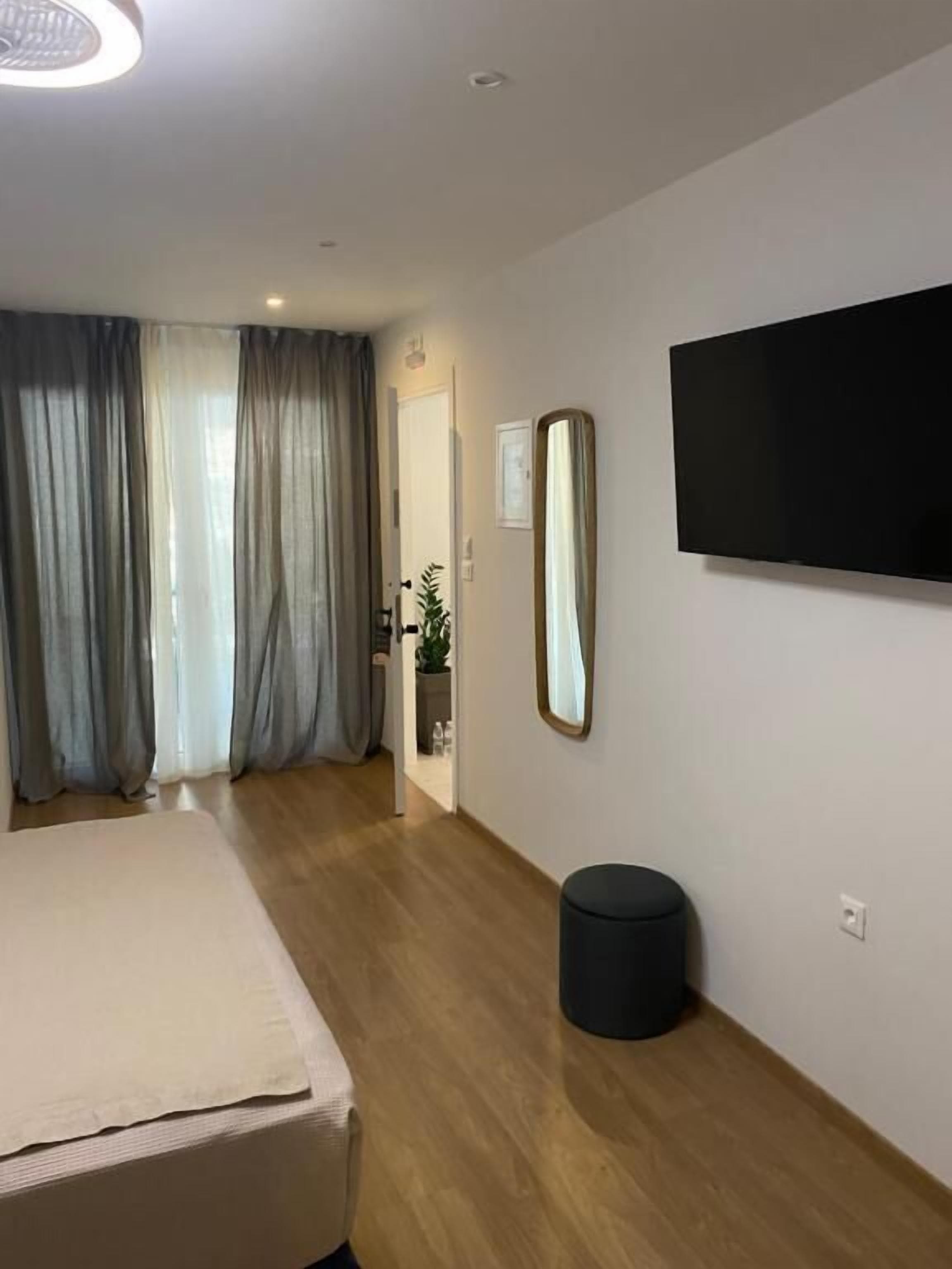 A modern hotel room with a bed, a flat-screen TV mounted on the wall, a round black trash bin, and a large mirror.