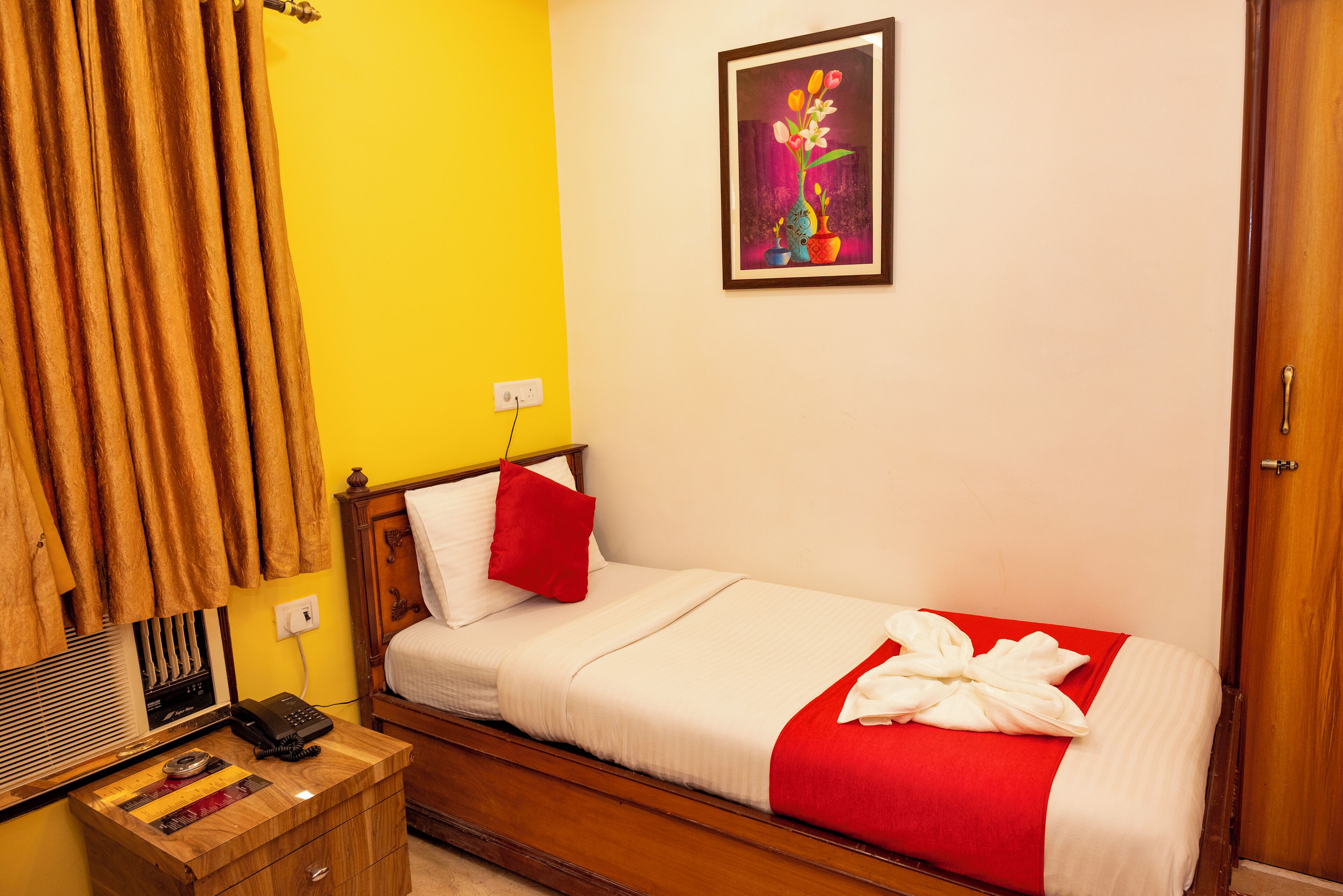 A single bed with a red and white bedspread, a wooden bedside table with a telephone, and a painting of flowers on the wall.